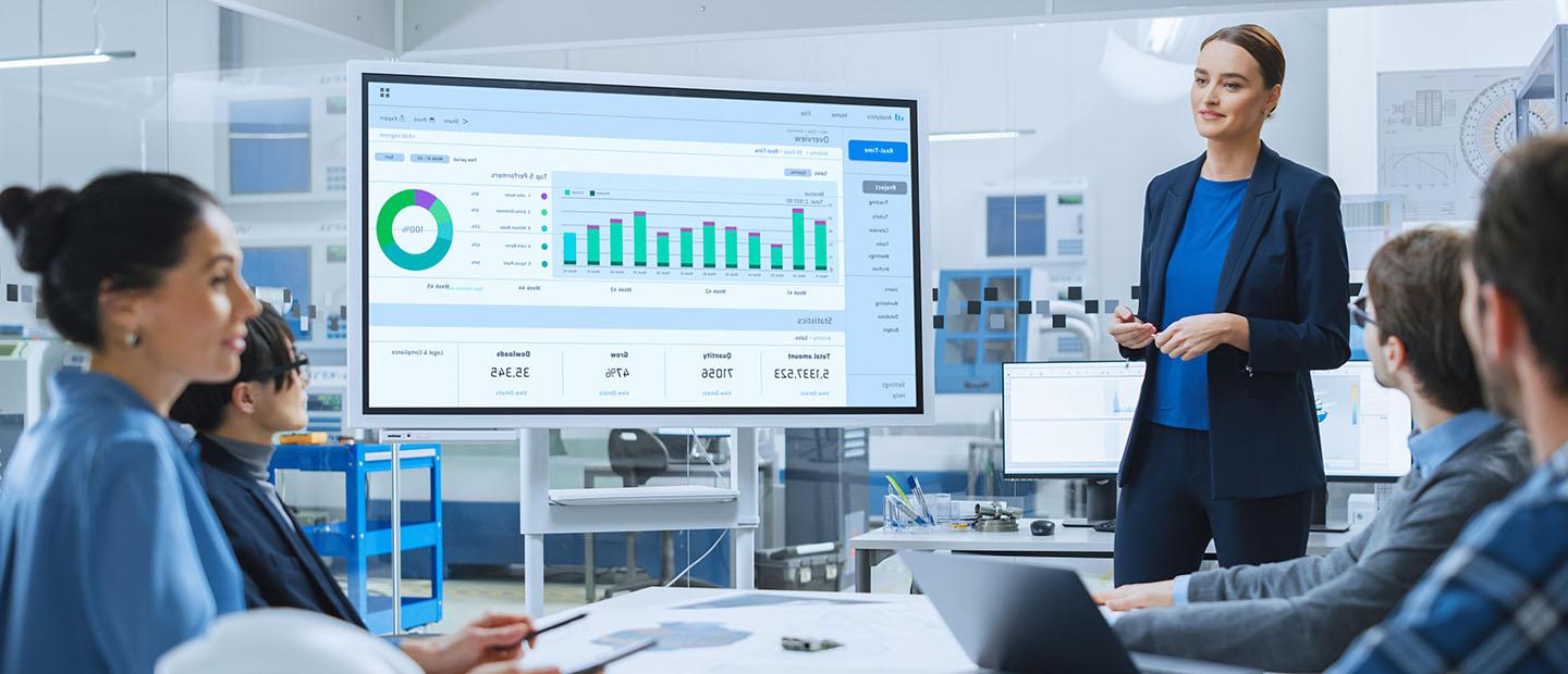 A woman presenting in front of a monitor displaying graphs.