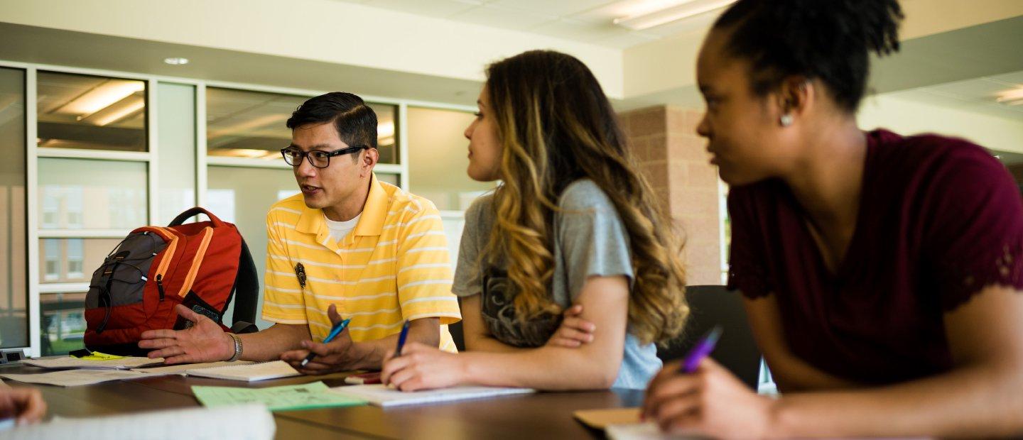 Two students sitting at a table look at a third who is speaking