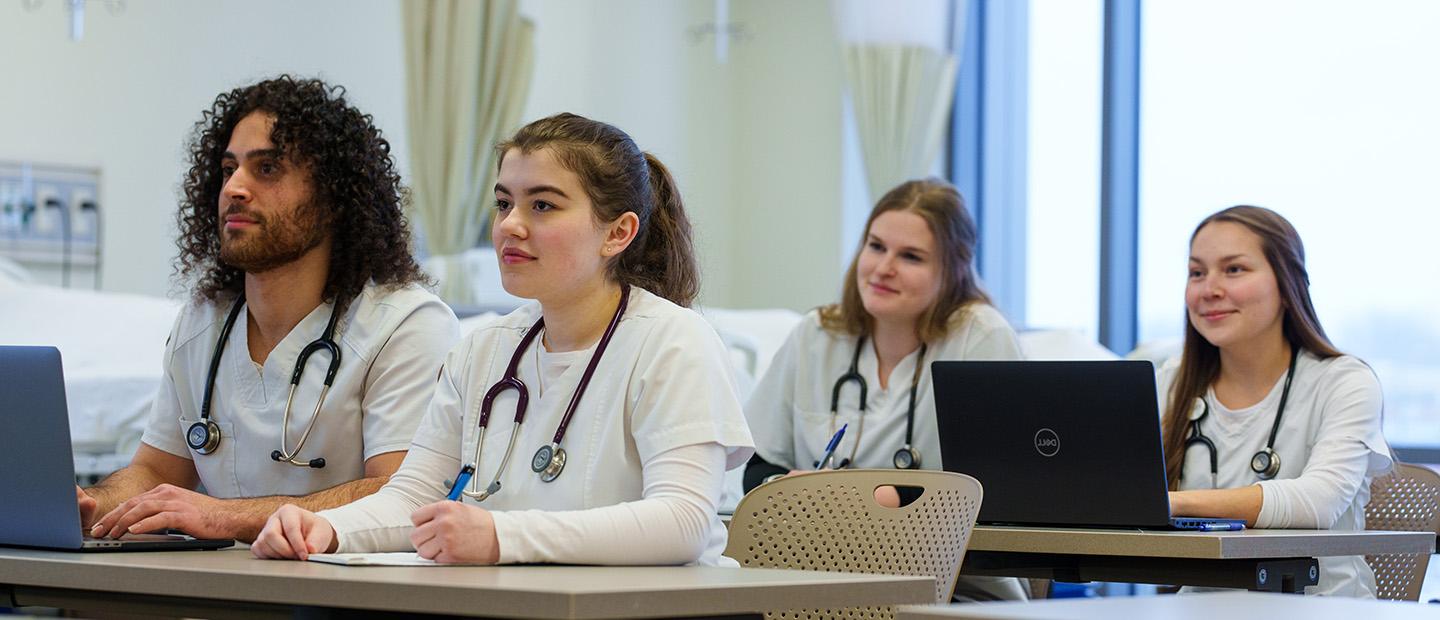 four students in white lab coats seated at a desk with stethoscopes